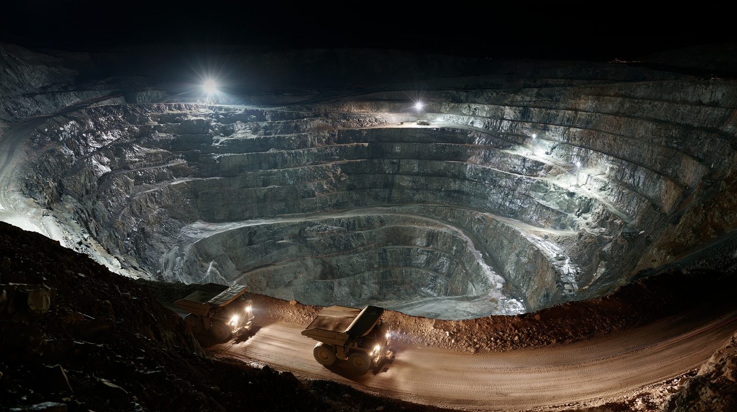 A deep terraced open-pit mine at night under floodlights, with two haul trucks on the road at its rim